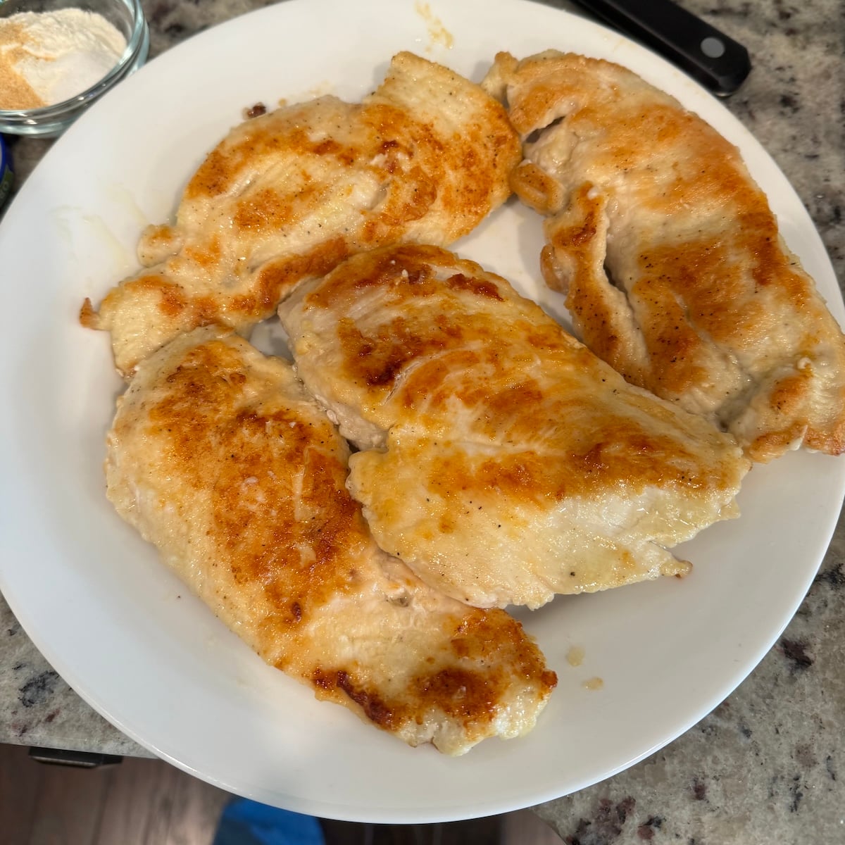 Four cooked, golden brown chicken breasts are arranged on a white plate placed on a granite countertop&mdash;perfect for adding to your favorite Creamy Chicken Pasta Recipe. A small bowl of flour and a knife are partially visible in the background.