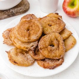 apple fritter rings piled on top of each other and drizzled with powder sugar on a white plate