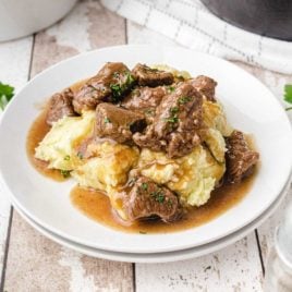 beef tips garnished with parsley and drizzled with gravy then served over mashed potatoes on a plate