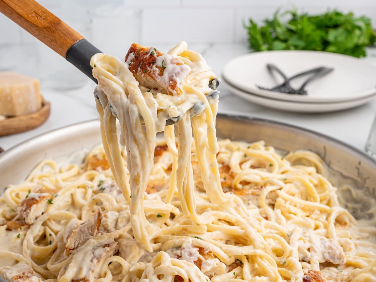 A serving spoon lifts creamy chicken pasta fettuccine Alfredo with pieces of grilled chicken from a pan, while plates and parsley are visible in the background&mdash;perfect for trying this creamy chicken pasta recipe at home.
