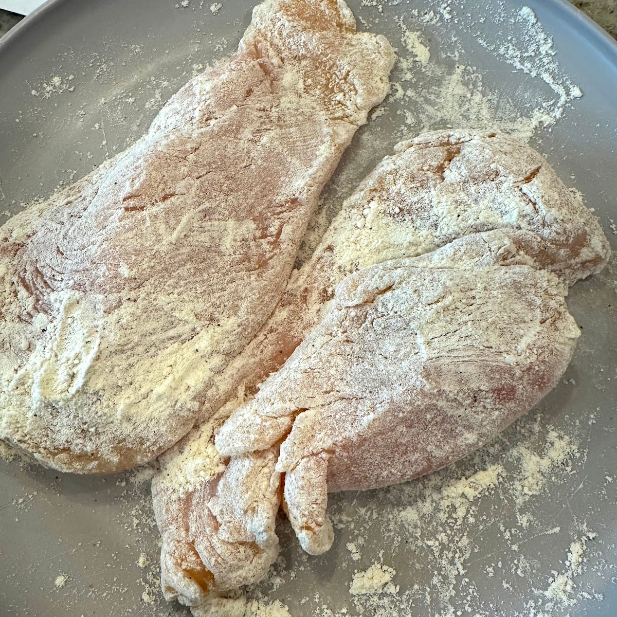 Two raw chicken cutlets coated in flour lie on a gray plate, prepped and ready to be transformed into a delicious Creamy Chicken Pasta recipe.