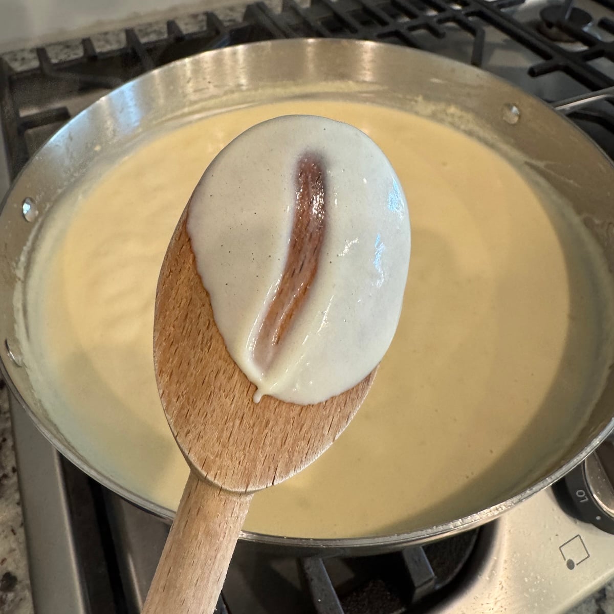 A wooden spoon coated with creamy white alfredo sauce is held above a pan filled with the same sauce, cooking on a stovetop&mdash;perfect for starting a Creamy Chicken Pasta Recipe. A clean line marks where a finger has run through the sauce on the spoon.