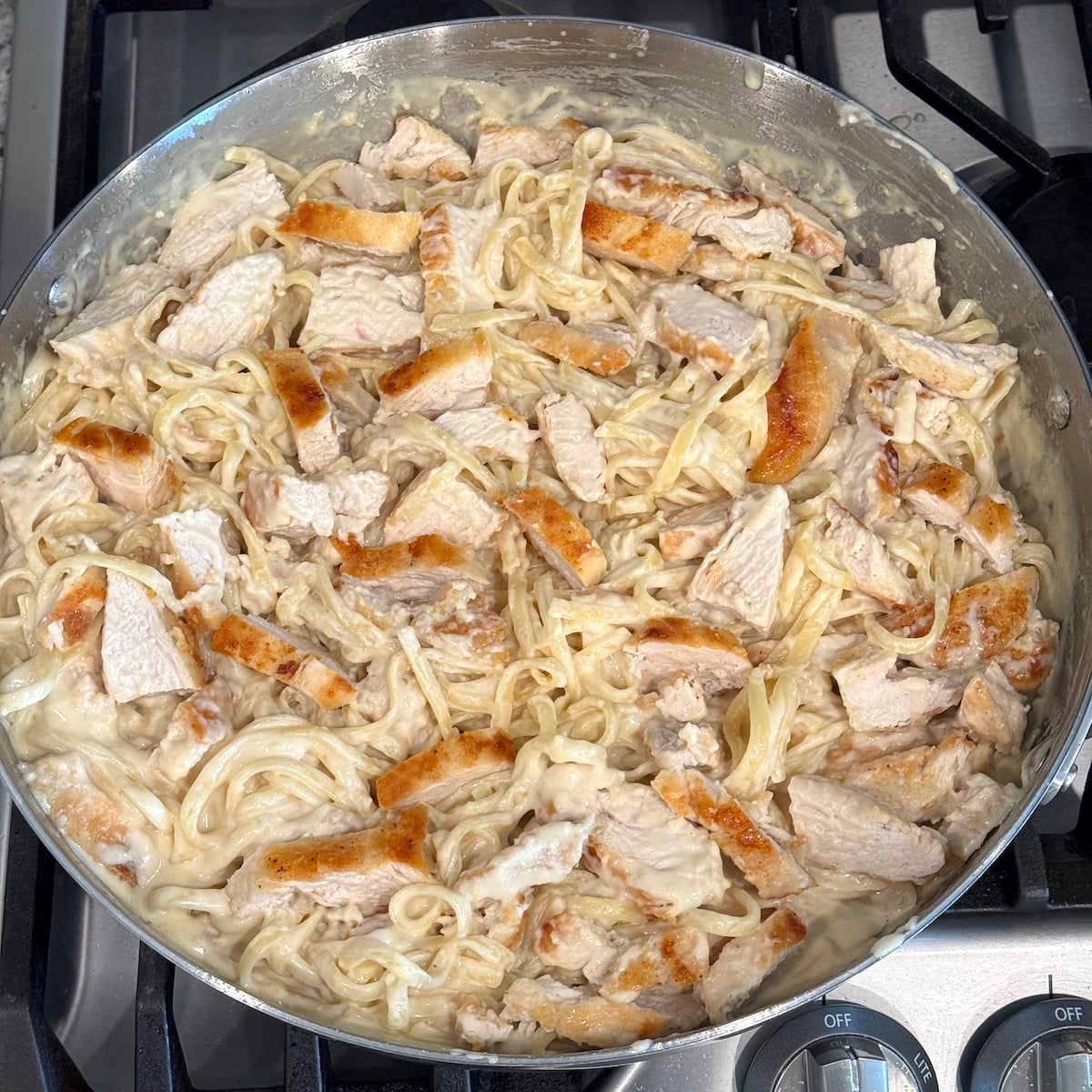 Four golden-seared chicken breasts, sliced and folded into creamy Parmesan linguine in a large stainless steel skillet on the stovetop.