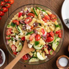 A bowl of Greek Pasta Salad with rotini, cherry tomatoes, cucumbers, red onions, olives, feta cheese, and fresh herbs. A wooden spoon rests in the bowl, surrounded by ingredients and empty plates on a rustic wooden table.