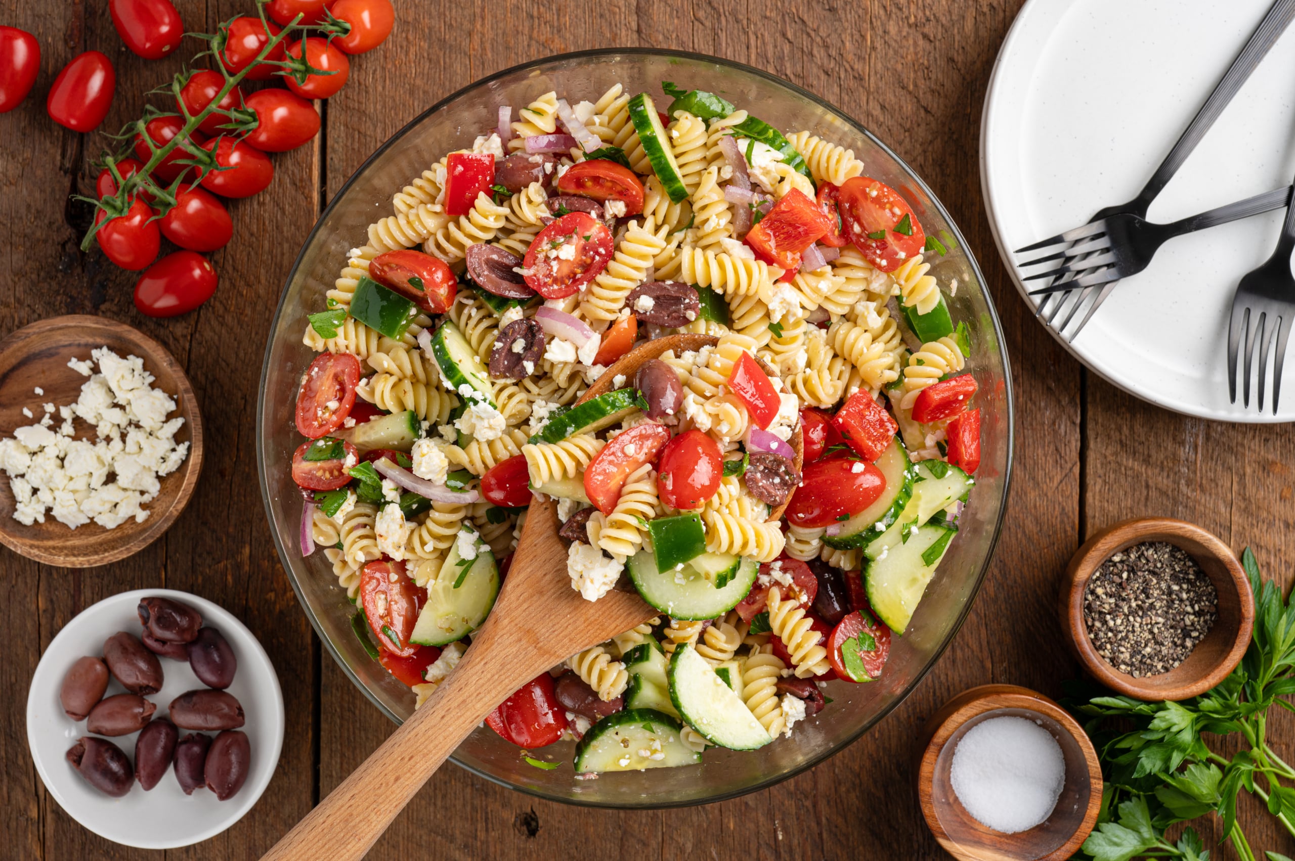 A bowl of Greek Pasta Salad with rotini, cherry tomatoes, cucumbers, red onions, olives, feta cheese, and fresh herbs. A wooden spoon rests in the bowl, surrounded by ingredients and empty plates on a rustic wooden table.
