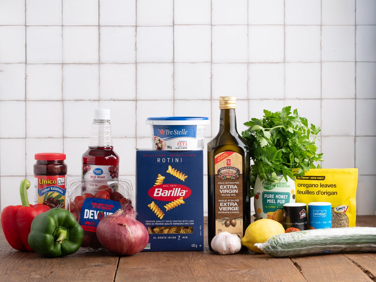 Assorted ingredients on a wooden surface, including everything you need for a fresh Greek Pasta Salad—bell peppers, red onion, cucumber, lemon, parsley, pasta, feta cheese, oregano, honey, kalamata olives, and more.