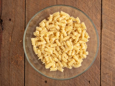 A glass bowl filled with uncooked rotini pasta sits on a rustic wooden surface, ready to become a delicious Greek Pasta Salad, viewed from above.