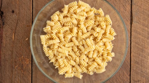 A glass bowl filled with uncooked rotini pasta sits on a rustic wooden surface, ready to become a delicious Greek Pasta Salad, viewed from above.