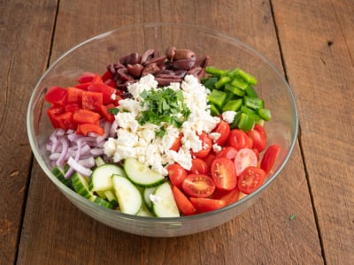 A glass bowl on a wooden table filled with chopped vegetables—perfect for a classic Greek Salad Recipe—with red bell pepper, kalamata olives, tomatoes, cucumber slices, red onion, feta cheese, and herbs.