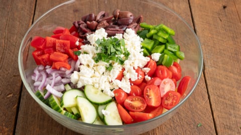 A glass bowl on a wooden table filled with chopped vegetables—perfect for a classic Greek Salad Recipe—with red bell pepper, kalamata olives, tomatoes, cucumber slices, red onion, feta cheese, and herbs.