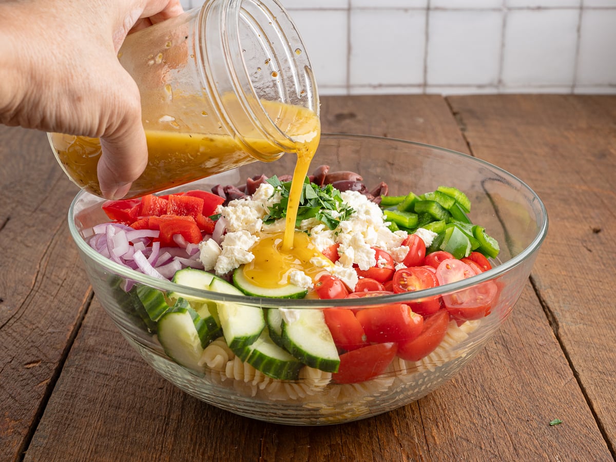 A hand pours vinaigrette dressing from a jar onto a bowl of Greek Pasta Salad with cucumbers, tomatoes, red onion, bell peppers, olives, and feta cheese on a wooden table. Try this Greek Salad Recipe for a fresh, flavorful meal.