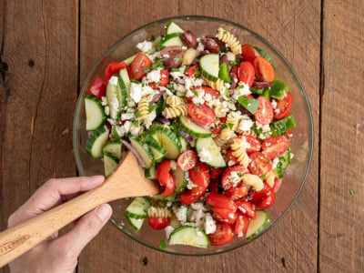 A hand holding a wooden spoon stirs a glass bowl filled with Greek Pasta Salad, featuring cherry tomatoes, cucumber slices, olives, feta cheese, and greens on a wooden table.