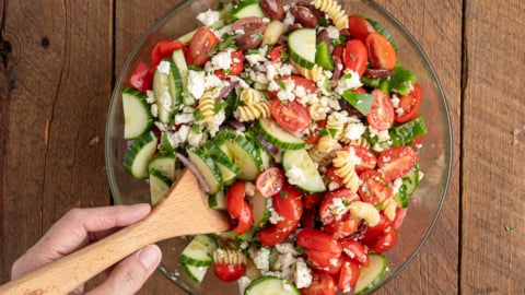 A hand holding a wooden spoon stirs a glass bowl filled with Greek Pasta Salad, featuring cherry tomatoes, cucumber slices, olives, feta cheese, and greens on a wooden table.