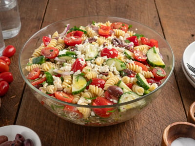 A glass bowl filled with Greek Pasta Salad featuring rotini pasta, cherry tomatoes, cucumber slices, red onion, Kalamata olives, feta cheese, and fresh herbs on a wooden table.