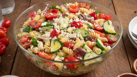 A glass bowl filled with Greek Pasta Salad featuring rotini pasta, cherry tomatoes, cucumber slices, red onion, Kalamata olives, feta cheese, and fresh herbs on a wooden table.