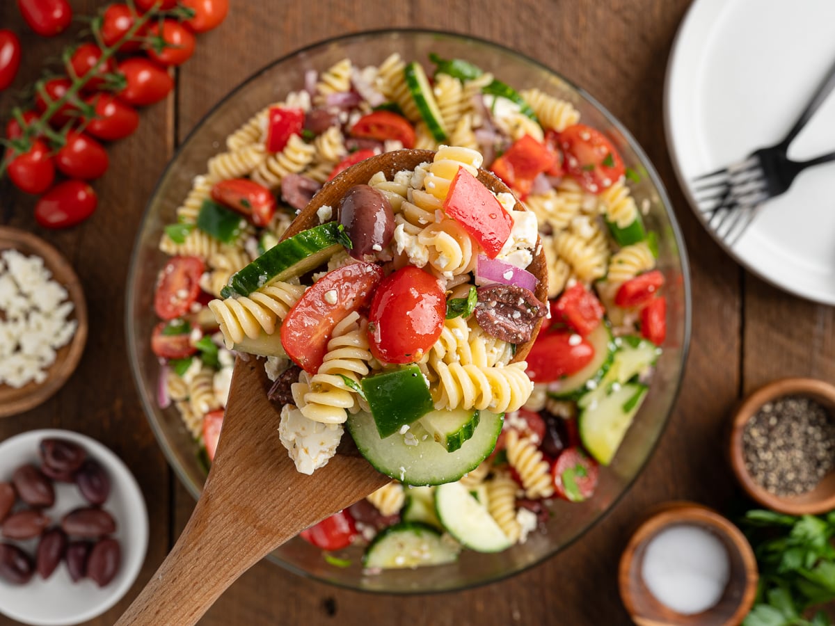 A wooden spoon holds a serving of Greek Pasta Salad with rotini, cherry tomatoes, cucumbers, olives, red onion, and feta over a large bowl. Surrounding the bowl are small dishes of ingredients for this classic Greek Salad Recipe.