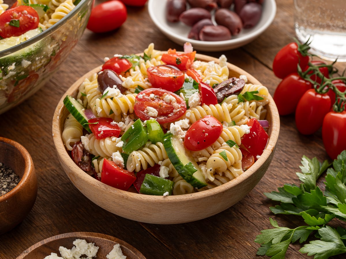 A wooden bowl filled with Greek Pasta Salad made of rotini, cherry tomatoes, cucumber, green bell pepper, Kalamata olives, red onion, and feta cheese sits on a wooden table with fresh herbs and vegetables nearby.