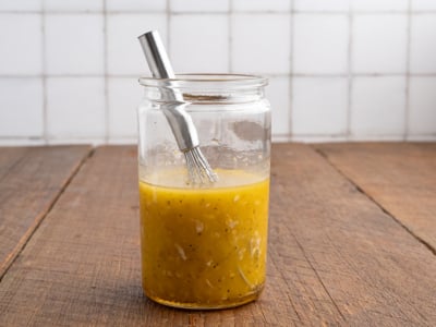 A glass jar filled with yellow salad dressing, herbs, and minced garlic—perfect for a Greek Salad Recipe—sits on a wooden table with a small metal whisk inside, set against a tiled white background.