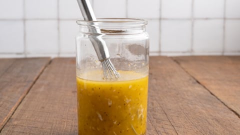 A glass jar filled with yellow salad dressing, herbs, and minced garlic—perfect for a Greek Salad Recipe—sits on a wooden table with a small metal whisk inside, set against a tiled white background.
