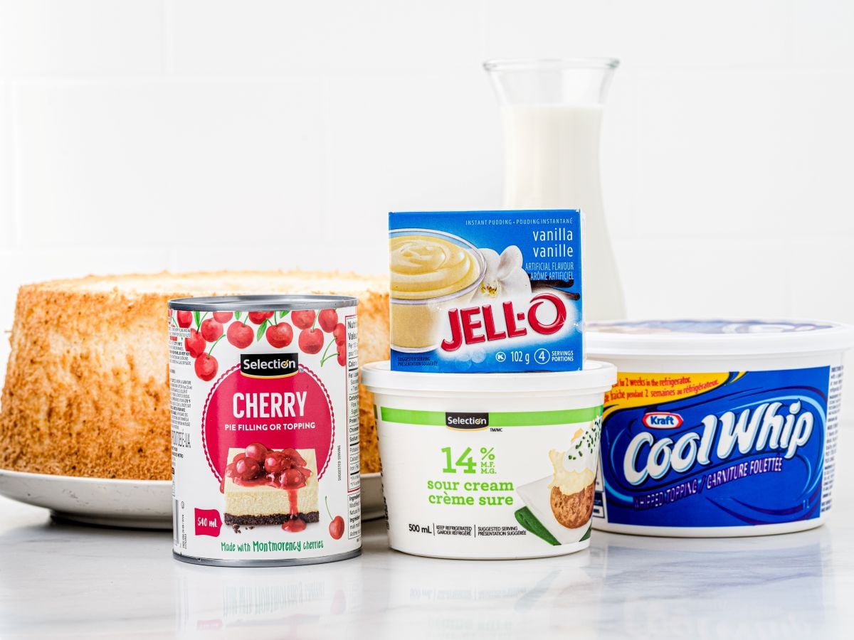 Various dessert ingredients on a counter, including cherry pie filling, vanilla Jello pudding mix, sour cream, Cool Whip, milk, and a plain sponge cakeโeverything you need for a delicious Heaven on Earth Cake Recipe.
