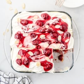 A glass baking dish filled with Heaven on Earth Cake, topped with whipped cream, cherry pie filling, and almond slices, with one corner piece missing. A plate with forks and a striped cloth are nearby.