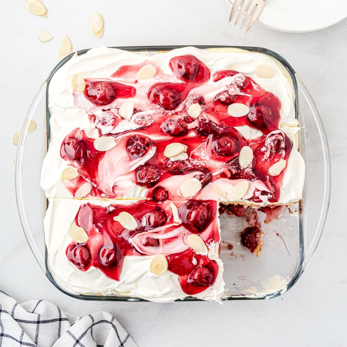 A glass baking dish filled with Heaven on Earth Cake, topped with whipped cream, cherry pie filling, and almond slices, with one corner piece missing. A plate with forks and a striped cloth are nearby.