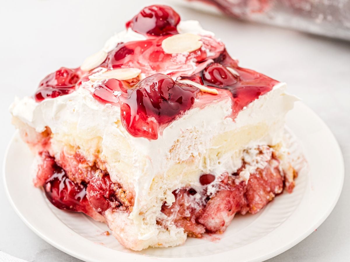 A slice of Heaven on Earth Cake on a white plate features whipped cream, cherry topping with whole cherries, and toasted almond slices, with layers of creamy and cake-like textures visible.