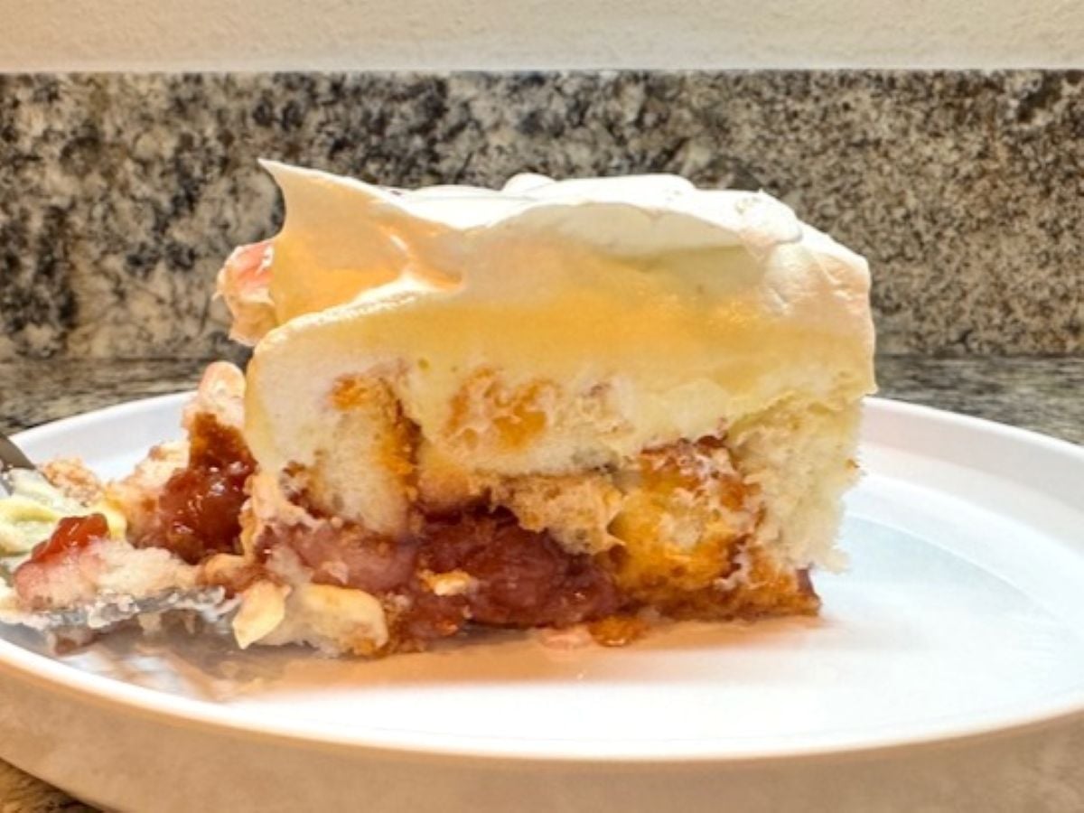 A slice of Heaven on Earth Cake with layers of whipped cream, custard, sponge cake, and fruit filling is served on a white plate against a granite countertop background.