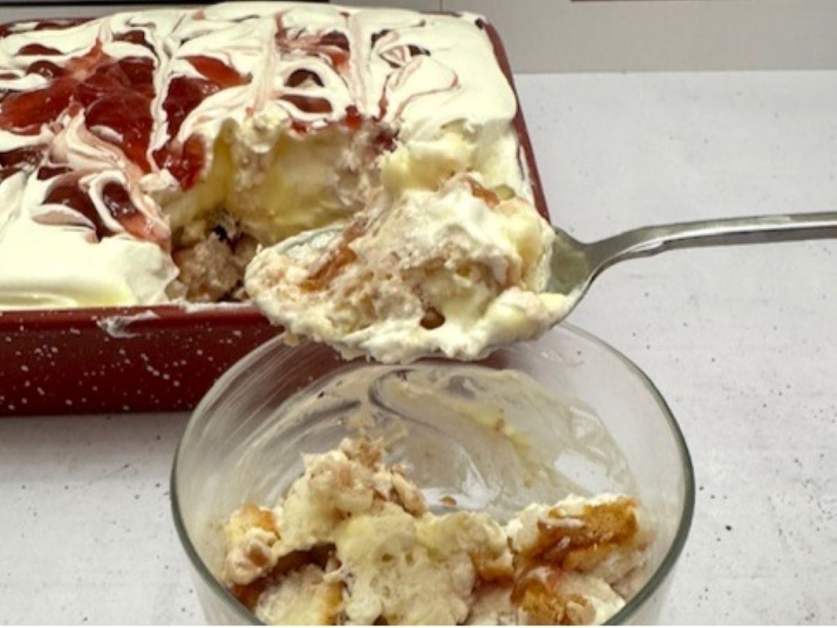 A glass dish of Heaven on Earth Cake topped with swirled red jelly sits on a table. A serving spoon scoops some into a clear bowl, revealing irresistible layers of cream, cake, and jelly.