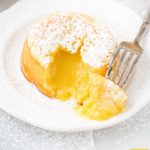 close up shot of lemon lava cake dusted with powdered sugar with a bite taken out showing its inside lemon layers on a white plate