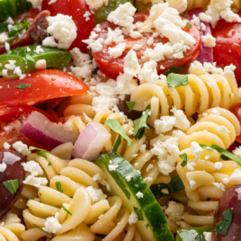 A close-up of Greek Pasta Salad with rotini noodles, cherry tomatoes, cucumber slices, red onion, Kalamata olives, parsley, and crumbled feta cheese. Text reads Greek Pasta Salad and vintage Greek Salad Recipe!.