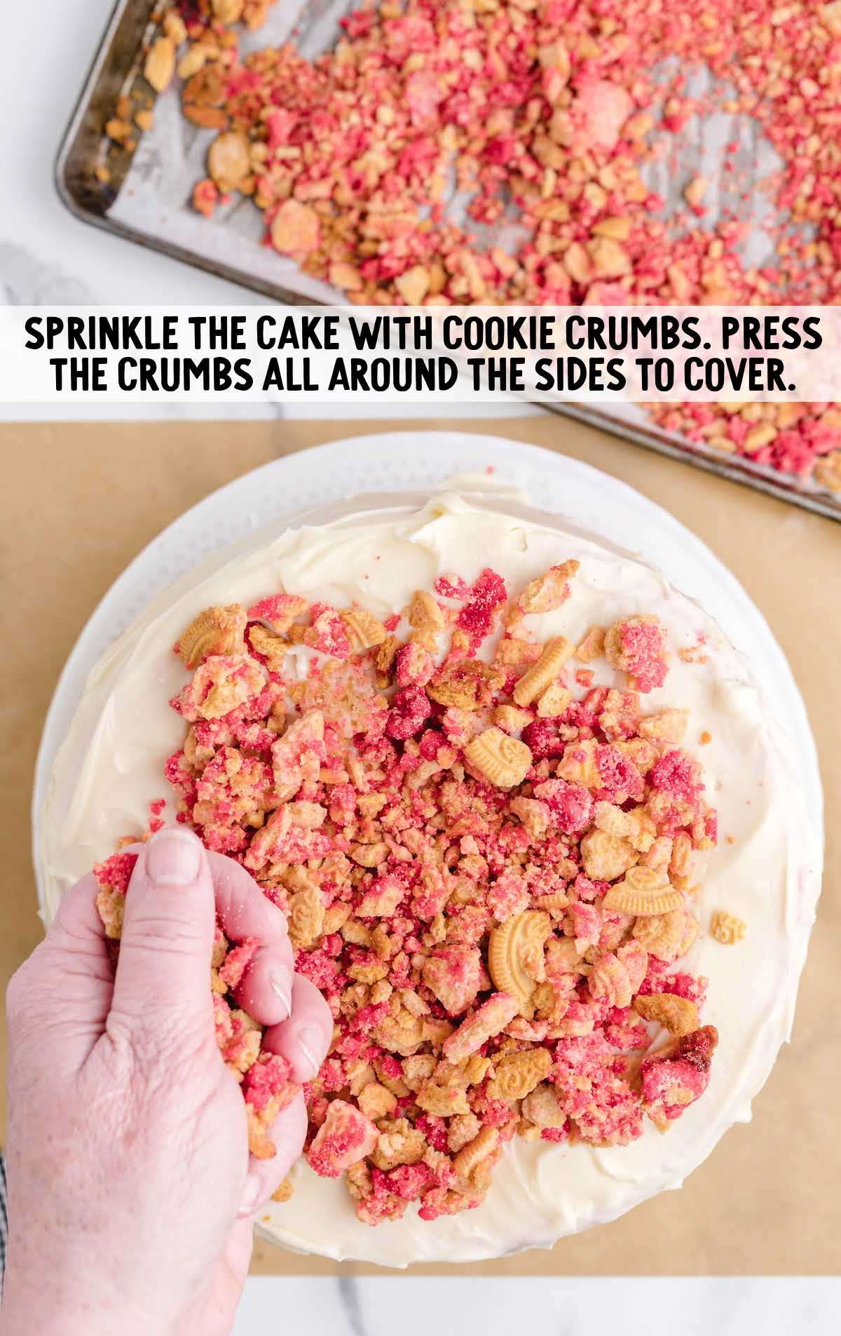 Hand pressing strawberry shortcake cookie crumbs onto a frosted cake to coat the top and sides.