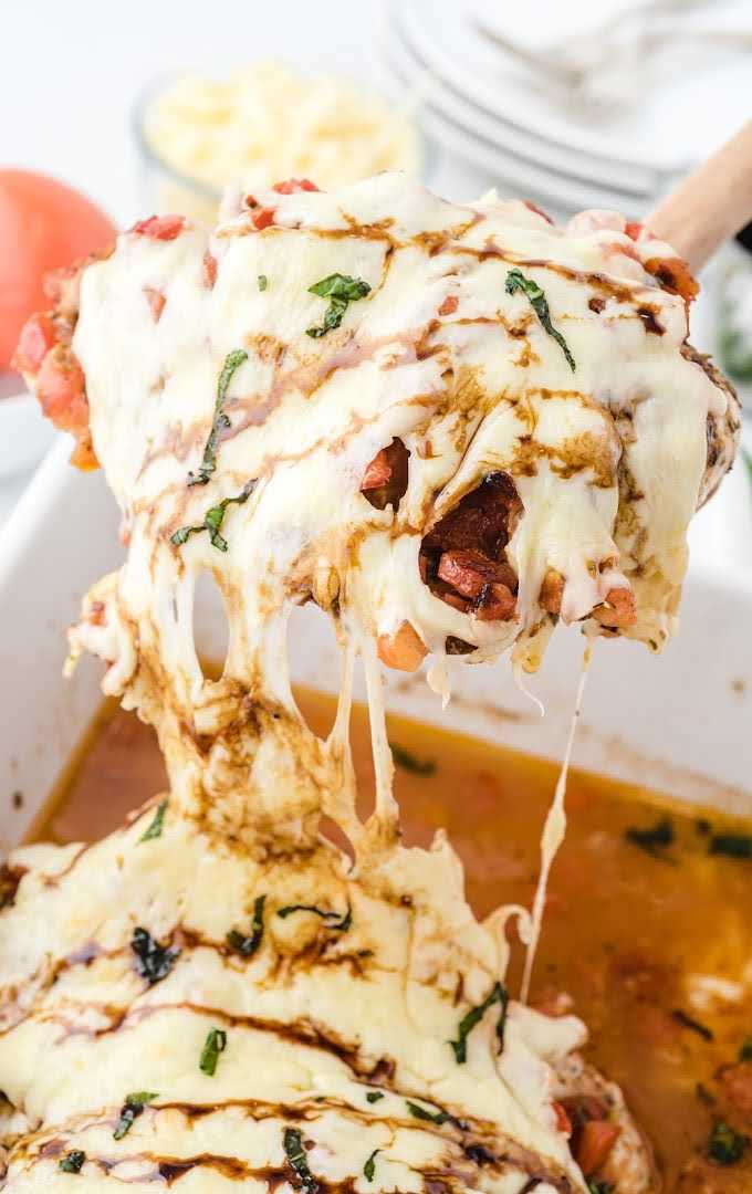 overhead shot of baked bruschetta chicken topped with mozzarella cheese then drizzled with balsamic glaze and basil being picked up with a wooden spoon
