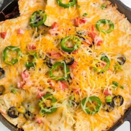 close up overhead shot of a skillet full of nachos topped with black olives, jalapeños, and tomatoes