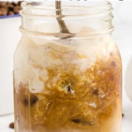 A mason jar filled with iced caramel macchiato, topped with whipped cream and caramel swirls, with a spoon inside. Text above and below reads: Iced Caramel Macchiato Recipe, Homemade Caramel Macchiato, Easy Recipe!.