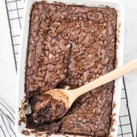 A pan of rich chocolate brownies, inspired by a classic Chocolate Dump Cake Recipe and topped with chocolate chips, sits partially scooped with a wooden spoon on a cooling rack.
