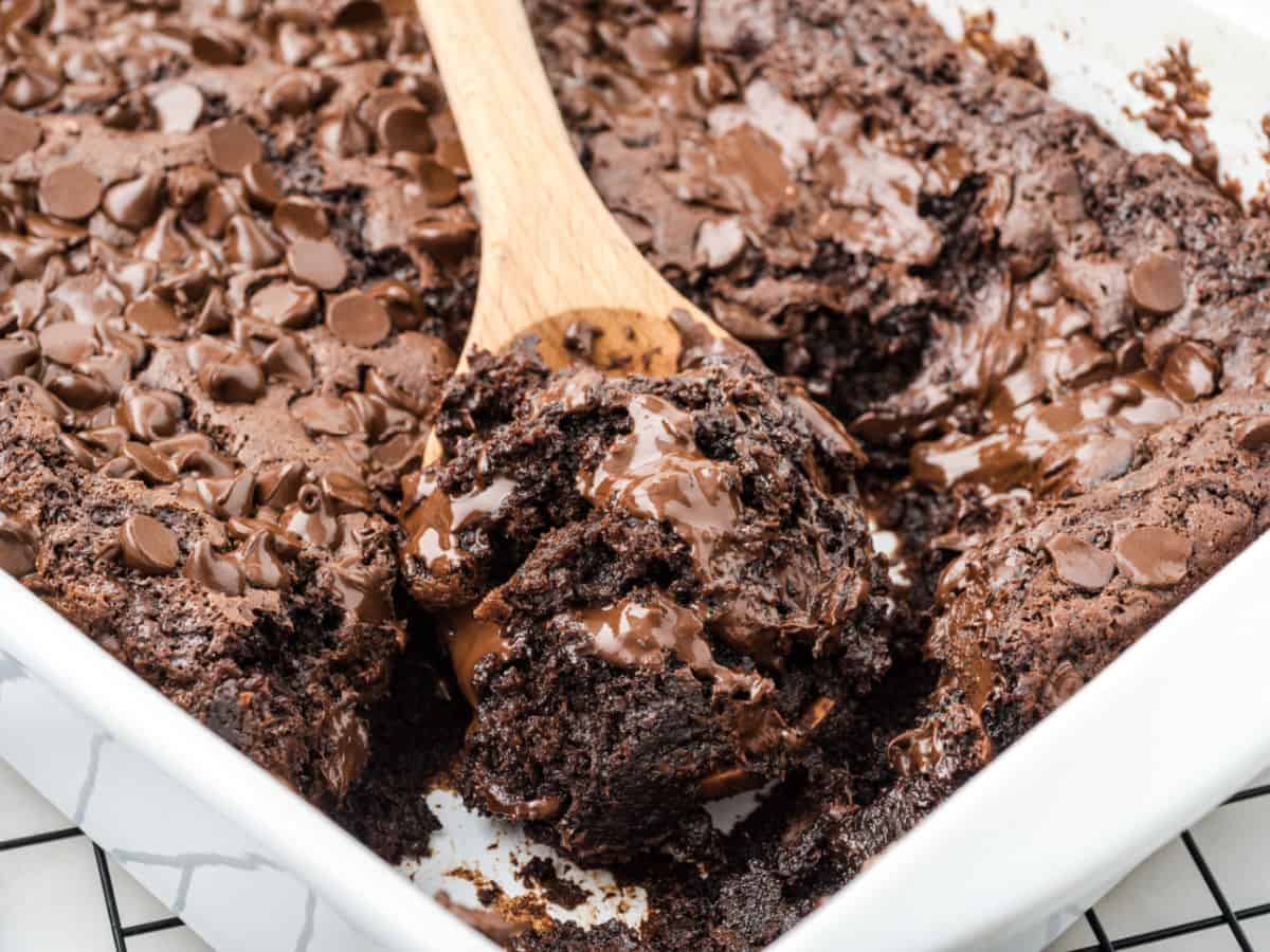 A wooden spoon scoops gooey, chocolate chip brownies from a white baking dish, reminiscent of a classic Chocolate Dump Cake Recipe. The brownies are rich, fudgy, and melted chocolate is visible throughout.