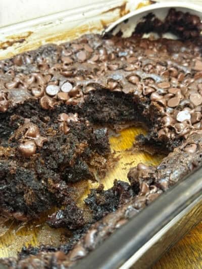 A partially eaten chocolate brownie in a glass baking dish, topped with melted chocolate chips and resembling a decadent Chocolate Dump Cake Recipe, shows off its gooey, rich interior. A spoon rests in the upper right corner.