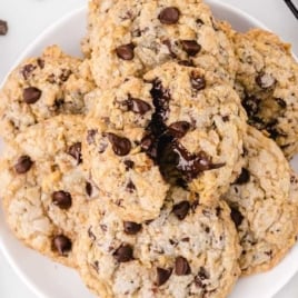 A plate of Doubletree Cookies with visible walnuts, surrounded by a bowl of chocolate chips, a black-and-white checkered cloth, and more cookies on a cooling rack.