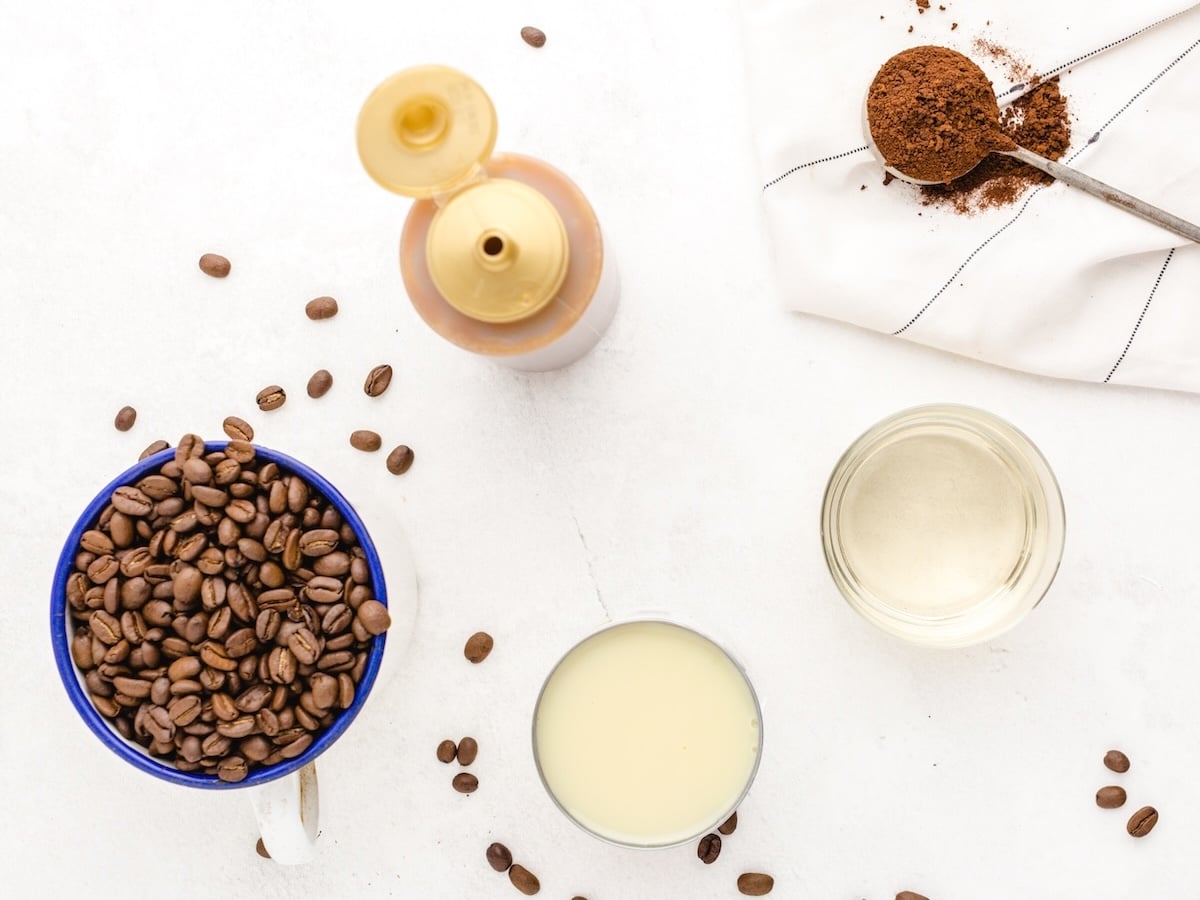 A mug filled with coffee beans, a glass of condensed milk, a jar of clear liquid, and ingredients for an Iced Caramel Macchiato Recipe are artfully arranged on a white surface, ready to make the perfect Iced Caramel Macchiato.