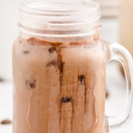 A mason jar mug filled with iced chocolate milk, featuring chocolate syrup swirls and ice cubes, evokes the look of an Iced Caramel Macchiato recipe against a light blurred background.
