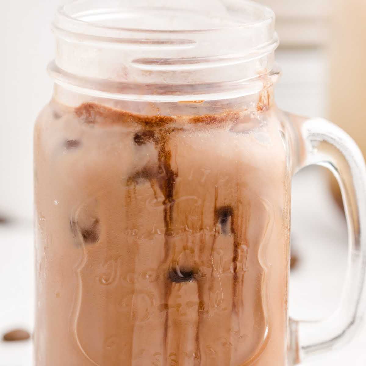 A mason jar mug filled with iced chocolate milk, featuring chocolate syrup swirls and ice cubes, evokes the look of an Iced Caramel Macchiato recipe against a light blurred background.