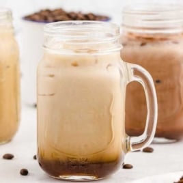 Four mason jars filled with iced coffee drinks, including an Iced Caramel Macchiato, sit on a white surface. Coffee beans are scattered around the jars, with a bowl of beans visible in the background. Perfect for trying any Iced Caramel Macchiato recipe!.