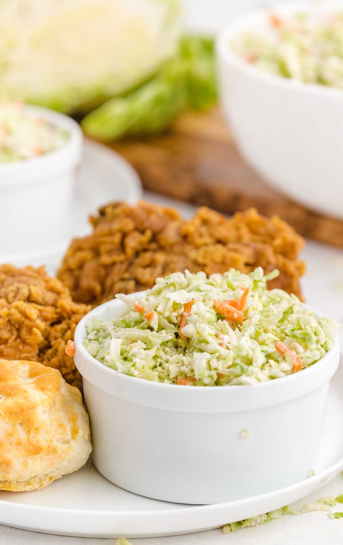 Bowl of creamy coleslaw placed on a white plate with fried chicken and a biscuit in the background.