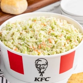 Large red and white bucket of creamy KFC-style coleslaw placed on a counter next to a biscuit and striped napkin.