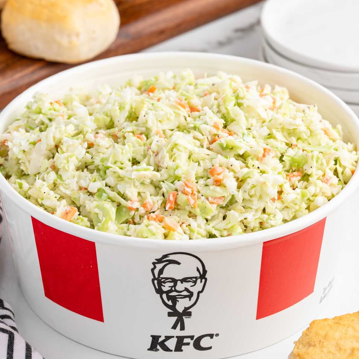Large red and white bucket of creamy KFC-style coleslaw placed on a counter next to a biscuit and striped napkin.