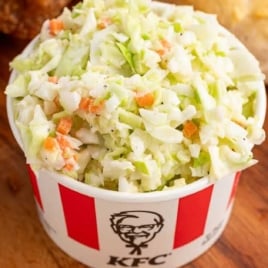 Red and white KFC cup filled with coleslaw on a wooden board next to fried chicken and a biscuit.
