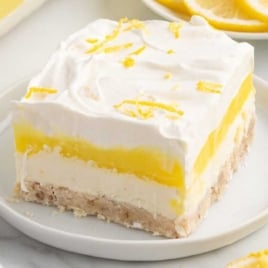 Slice of lemon lush dessert on a plate garnished with lemon zest, with whole lemons and sliced lemons in the background.