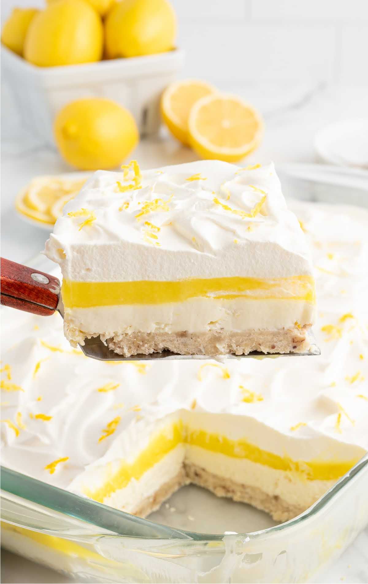 Full slice of lemon lush dessert on a plate topped with lemon zest, surrounded by lemon slices and a dish of zest.