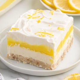 Slice of lemon lush being lifted from a glass dish with creamy layers of pudding and whipped topping, and a bowl of lemons in the background.